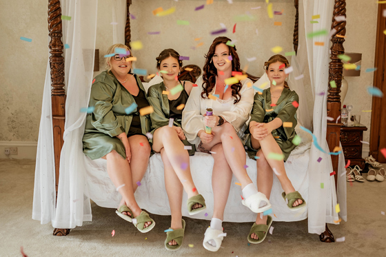 Bride and her bridesmaids do confetti shot sitting on four poster bed